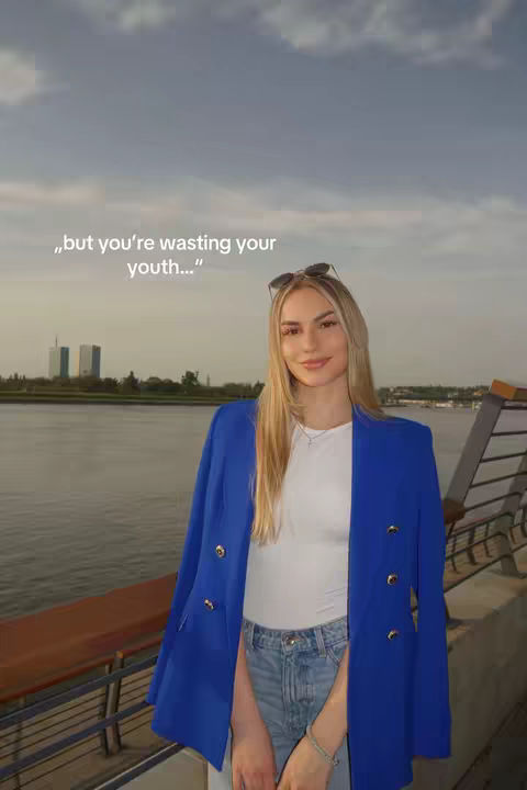 A woman with long blonde hair is standing by a body of water, wearing a white top, light blue jeans, a blue blazer and sunglasses on top of her head. There are buildings visible in the distance and the sky is cloudy.