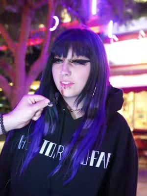 A medium shot shows a young woman with black hair with streaks of purple, wearing a black hoodie, and a hand holding a microphone. The background is blurred with trees and neon lights.