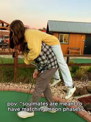 A woman is hugging a person from behind, they are both on what seems to be a mini golf course. The woman is wearing a yellow sweater, while the person being hugged is wearing a plaid shirt and grey pants. In the background, there are wood buildings and trees with an appealing environment.