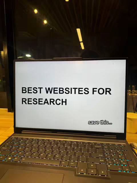 A laptop displaying a screen with the text "BEST WEBSITES FOR RESEARCH" in bold sans-serif font. Below the text, smaller text reads "save this...". The laptop is open on a desk, and the surrounding environment is dimly lit with warm, yellow lights.