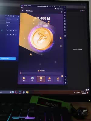 A person is showing a computer screen that is running an app. The app is in the middle of the screen and looks like some sort of crypto-currency or trading platform that displays a coin with the letter "T" in the middle with a gold color, as well as a count of 7.400 M. The screen itself is on a black monitor, with a dark background behind. The app seems to be running in an emulator. In the front of the image, a keyboard is seen with the letters of the keyboard visible and a neon green and black key chain that appears to be Honda.