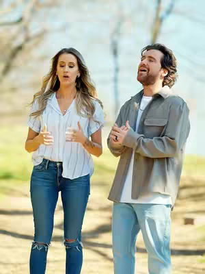 The cover image features two individuals in a natural outdoor setting. A woman and a man both appear to be mid-sentence with their mouths open, suggesting they are singing or speaking. The man is on the right, wearing a jacket, while the woman is on the left, wearing a shirt. The background is blurred, suggesting a shallow depth of field, with a bright sky.