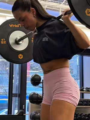 A woman in a gym lifting weights, wearing athletic apparel. The image is taken from a side angle, showing the woman's face in profile and the weight bar on her shoulders. She is focused on the exercise. The lighting suggests an indoor gym environment. The color palette includes dark colors of the weight bar and the black top, blending with pink shorts and the skin tone of the woman. The background has blurred elements of the gym.