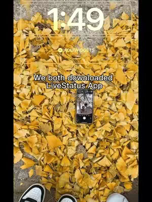 The cover image showcases a close-up view of fallen yellow leaves, creating a textured, autumnal background. In the center, there is a smartphone lying on the leaves, displaying a photo of two people. Overlaying the scene is the time '1:49'. Above the phone, text reads: 'We both downloaded LiveStatus App.' Below, the lower portions of two pairs of shoes are visible, suggesting the perspective is from someone standing above the leaves.