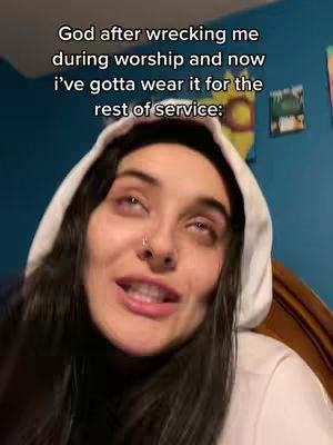 A selfie-style video cover image. A young woman with long, dark hair and a white hoodie. She is looking upwards with her eyes closed slightly and a subtle expression on her face. Text overlay reads: 'God after wrecking me during worship and now i've gotta wear it for the rest of service:' The background includes hints of a blue wall and a bed frame. The overall composition is a close-up, emphasizing the woman's face and emotional reaction.