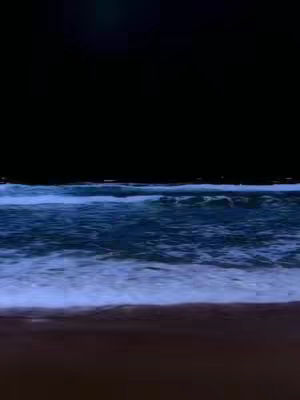 A nighttime beach scene. Dark black covers the top half, while the bottom half reveals a shoreline. The waves near the shore are visible, with white foam breaking against the sand. There's a gradient of dark colors fading upwards.