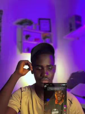 A man with dark skin and a black cap is in the foreground, holding his right hand to his head. He appears to be in a well-lit room with purple lighting, against a backdrop of a wall with a shelf and framed picture. In the bottom right, there's evidence of a mobile phone screen displaying another person.