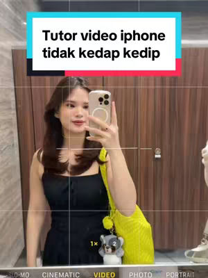 A woman is in a bathroom, taking a mirror selfie with an iPhone. She wears a black dress and holds a yellow bag. Overlay text informs the video will teach iphone tips.