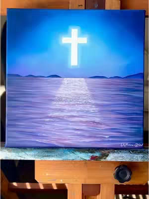 A painted canvas on an easel depicting a white cross emitting light in the sky that directs light towards a water surface. The water surface with purple and violet hues, and in the distance, dark blue mountains. The painting is secured on a wooden easel.
