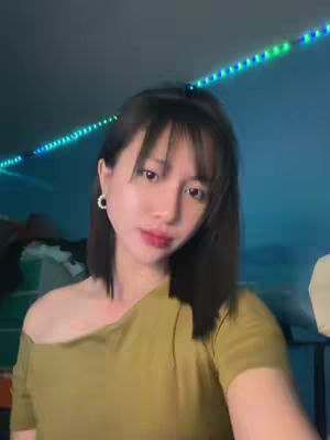 A young woman with dark hair and bangs poses for a selfie. She wears a yellow off-the-shoulder top and has a simple earring and a slight smile. The background is out of focus, but displays a gradient of green and blue lights.