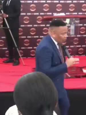A man in a blue suit is speaking into a microphone on a stage with a red carpet. Another person in a black suit and tie is standing to the man's left, and the blurred heads of audience members are visible. The background has a pattern of connected circles.
