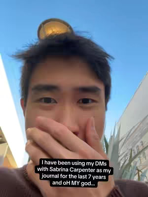 A young man, seen from the chest up, is the focus of the image, his hand covering his mouth in a gesture of surprise or embarrassment. He's looking directly at the camera with dark hair and skin, and a small golden object perched on top of his head. The background is a blue sky with hints of what appears to be a building.
