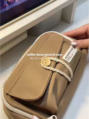 The image showcases a close-up of a tan, coffee bean colored pencil case. The case is rectangular and secured with a strap and a wooden button. It appears to be resting on a light-colored surface and a hand with painted nails is holding the case. In the background, there's a glimpse of a light-colored shelf with books or similar items placed on it. There is text overlayed on top, that reads, 'coffee bean pencil case @tiemnhochu.do' and another white label which reads 'my study accept'.