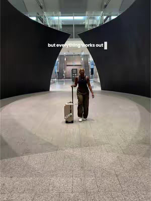 A woman walks towards the camera in a large, modern hall; her white suitcase is pulled along. The hall has high ceilings, a polished floor and dark, angular structural elements. Text overlay, "but everything works out", is present.