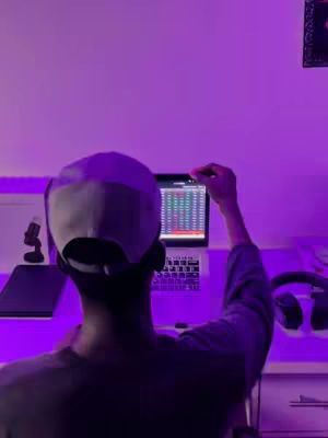 A person wearing a cap is sitting in a room with purple lighting, facing away from the camera and looking at a laptop. The laptop screen displays a chart.