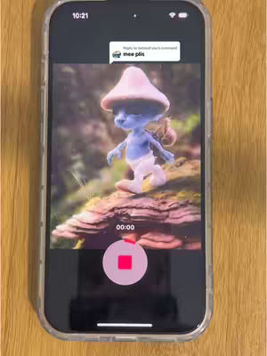 A smartphone displaying a video screen. The screen shows a cartoon Smurf character in a forest setting. The phone is encased in a clear case and rests against a wooden surface. Overlaying is a comment from other user.