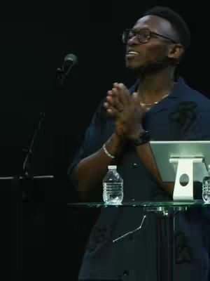 A man in a dark blue shirt and glasses stands on a stage with his hands clasped in front of him, as if in prayer. A microphone and a water bottle rest on a table in front of him.