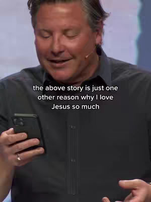 A man with short, graying hair is holding a black smartphone in his left hand. He is wearing a dark gray button-down shirt and a gold ring on his ring finger. The background is a blurred mix of colors, likely a stage or studio setting. Text overlayed on the image reads: 'the above story is just one other reason why I love Jesus so much'.