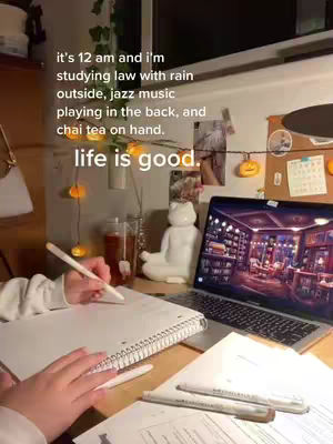 A well-lit shot of a desk setup. A laptop displays a dimly lit room, a notebook is open with a hand holding a pen. There is a glass of tea, a cat figurine, and fairy lights adorning the desk. The composition suggests a cozy and focused study environment.