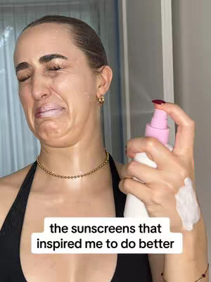 A woman is shown with sunscreen on her face in an image, with her eyes squeezed shut and mouth slightly open. She appears to be reacting to being sprayed with the sunscreen spray from a pink and white bottle. The background includes what looks like a shower or tub and a light-colored wall.