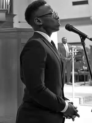 A black and white photo shows a man with short, dark hair and glasses speaking into a microphone. He is wearing a dark suit and tie, and his expression is serious. The background is a blurred interior space that appears to be a church or performance hall.