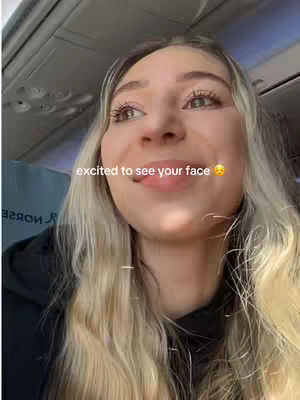 A selfie of a woman inside an airplane, looking upwards with an excited expression. The woman has long, blonde hair, and the background reveals the interior of an airplane.