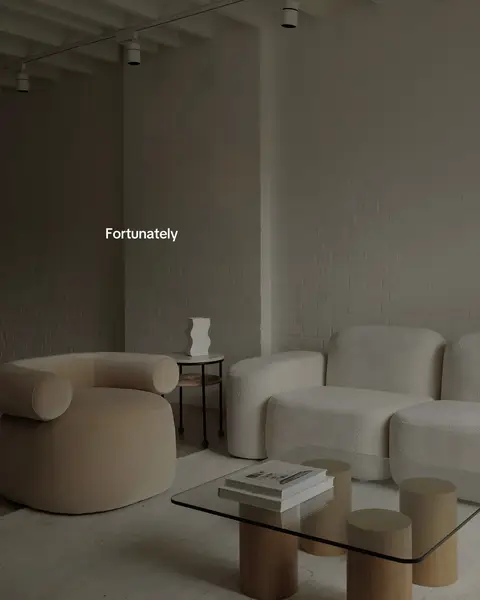 A minimalist living room scene with a beige, curved armchair and a white, rounded sofa. A glass-top coffee table sits on light-colored wooden legs, holding a stack of books. A small side table with a white vase is next to the sofa. The wall is white with track lighting.