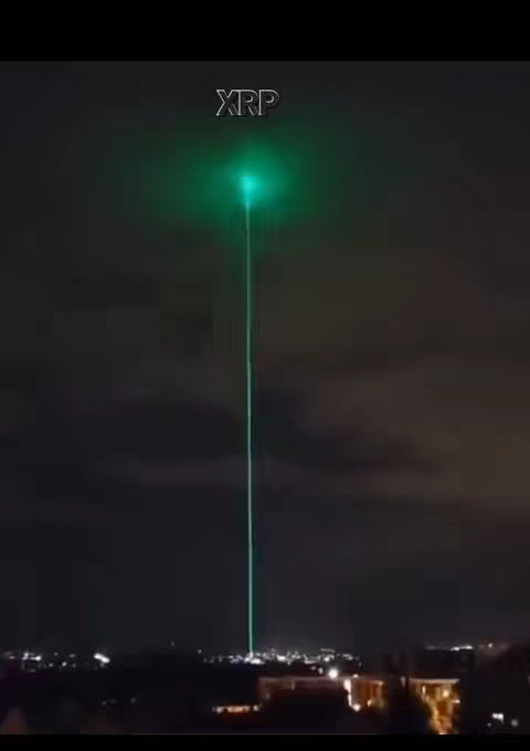 A nighttime view shows a city skyline under a dark, clouded sky. A bright green beam of light shoots straight upwards from the city, potentially a laser or visual effect, with the word "XRP" overlaid near the top.