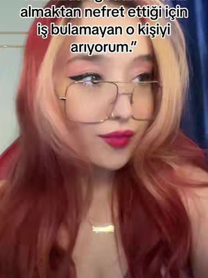 The image is a selfie of a person with colorful hair, glasses, and bright lipstick, looking toward the camera. Text overlays the image in the top-left corner. The background seems to be a room with a blue curtain visible.