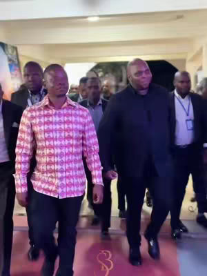 The image shows a group of people walking in a well-lit hallway. In the foreground are two men, one wearing a patterned pink and white shirt and the other in a black suit. A red carpet with a decorative emblem is underfoot, and several other people, primarily men in suits, walk behind them. The lighting is bright, and the overall impression is of a formal event or gathering.
