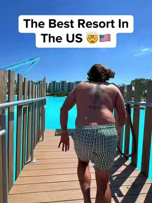 A man with short-cropped hair walks on a wooden boardwalk, with a turquoise lagoon and distant buildings in the background under a blue sky. Text overlays at the top, with a face with steam emoji and a US flag.