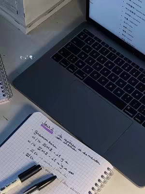 A top-down shot of a desk with a laptop and notepad. The laptop is partially open, showing a screen with a Korean language lesson. A notepad with handwritten notes in both French and Korean lies open beside it, along with two pens. The lighting is soft and natural, suggesting an indoor setting. The overall aesthetic is clean and organized with a study-focused feel.