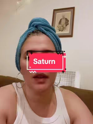 The cover image features a woman centered in the frame, wearing a blue towel wrapped on her head like a turban. A red rectangular text box with the word "Saturn" is placed over her face. She wears hoop earrings and a white tank top. The background includes a wall, a framed picture, and a small white appliance.