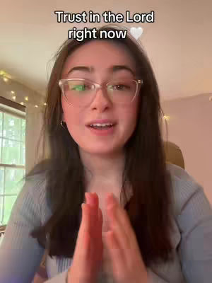 A young woman with long brown hair and glasses is in a room with a window and Christmas lights in the background. She is smiling at the camera with her hands clasped in front of her.