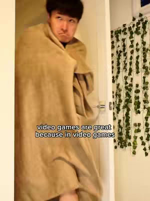 The image shows a man wrapped in a blanket, peeking out from behind it with a slightly displeased expression. The background includes a door and greenery along the wall, hinting at a domestic setting. Text overlay at the bottom reads, 'video games are great because in video games'.