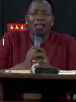 The image shows a woman with closed eyes holding a microphone, seemingly singing or speaking with emotion. She is dressed in a maroon top, and the background is a dark setting. Overlaid on the image are three yellow hand-praying emojis on a red background.