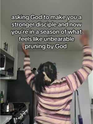 A person with dark hair wearing a striped sweater is standing in a kitchen with their arms raised, presumably in a celebratory or worshipful gesture. Text overlayed indicates religious themes.