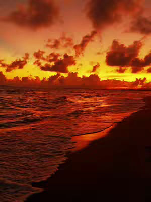 A photograph capturing a sunset over the ocean. The sky is filled with orange and yellow hues, with dark, cloudy formations scattered throughout. The sea reflects the colors of the sky, appearing predominantly orange. The shoreline is visible on the right, with a dark silhouette against the bright sky. The lighting is dramatic, casting the scene in warm tones.