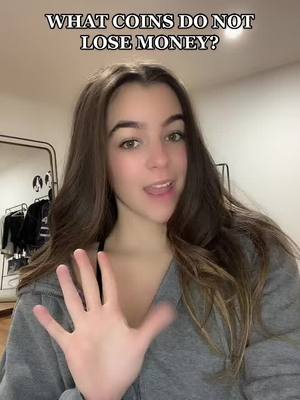 A woman with long brown hair is centered, looking at the camera and speaking. Behind her is a blurry room, and text overlay reads, 'WHAT COINS DO NOT LOSE MONEY?'. She holds up her right hand showing five fingers.
