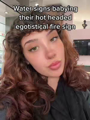A medium shot features a young woman with long, wavy auburn hair. She has bright green eyes, a small nose ring, and is wearing a dark top. The overlay on the image reads, 'Water signs babying their hot headed egotistical fire sign'.