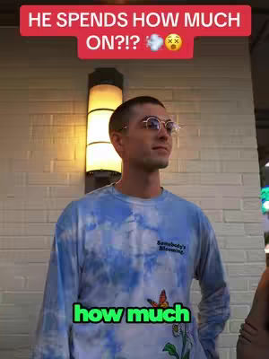 A man wearing glasses and a blue tie-dye shirt stands in front of a brick wall with a bright light fixture.