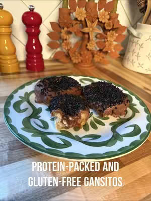 The image showcases a plate with three chocolate-covered pastries, sprinkled with dark flecks, presented on a surface with wooden textures. The plate has a white base decorated with a green pattern. The background displays additional culinary elements such as spice grinders with red tops, and a decorative wooden piece. The text overlayed reads 'PROTEIN-PACKED AND GLUTEN-FREE GANSITOS'.