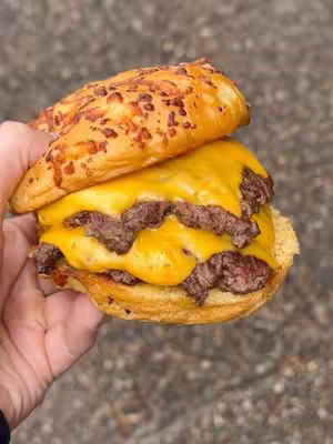 Close-up shot of a hand holding a double cheeseburger. The burger consists of a sesame seed bun, two beef patties covered in melted yellow cheese, and a layer of cheese in between. The background is blurred, showing an out-of-focus gray surface.