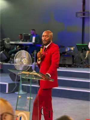 A man in a red suit stands on a stage holding a microphone and a book. The background shows a large screen and some stage equipment.