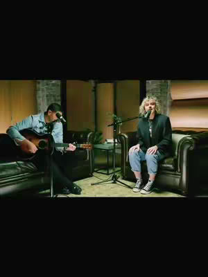 The image shows two people in what looks like a recording studio. A man on the left playing a guitar and a person sitting on the right with a microphone.