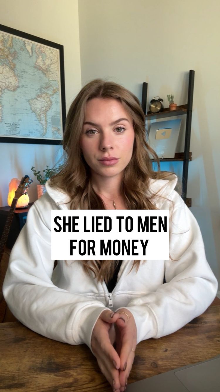 A woman in a white hoodie is seated at a wooden desk, looking directly at the camera with a neutral expression. A text overlay stating, "SHE LIED TO MEN FOR MONEY" covers a portion of the image. The background includes a framed world map, a wooden shelf, and a lamp, indicating a home or office setting. The composition is well-lit, with a focus on the speaker.