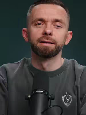 The image is a medium shot of a man with a short beard and short hair, facing the camera. He is wearing a dark gray long-sleeved shirt. In front of him is a black microphone. The background is a solid, muted green color. The lighting appears to be soft and even.
