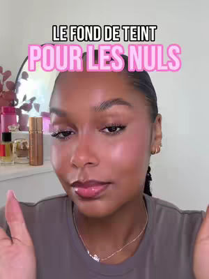 A close-up shot of a woman with flawless makeup. Text overlay in a bubble reads 'Le fond de teint pour les nuls' in a pink bubble with a white outline. She is framed from the chest up with her hands gesturing downwards.