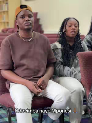 The cover image features two individuals, a man and a woman. The man is in the foreground, wearing a brown shirt, a gold chain and a cap. The woman is seated next to him, dressed in a gray jacket. They are both seated in a setting with red chairs. The text overlayed is at the bottom.