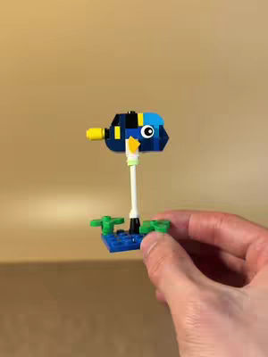 A hand holds a Lego creation of a blue fish on a white stick. The fish has different colored blocks and a large eye. The background is a plain tan color.